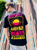 Person wearing a black t-shirt with 'Sunset Sessions' design on the back, standing against a colorful wall.