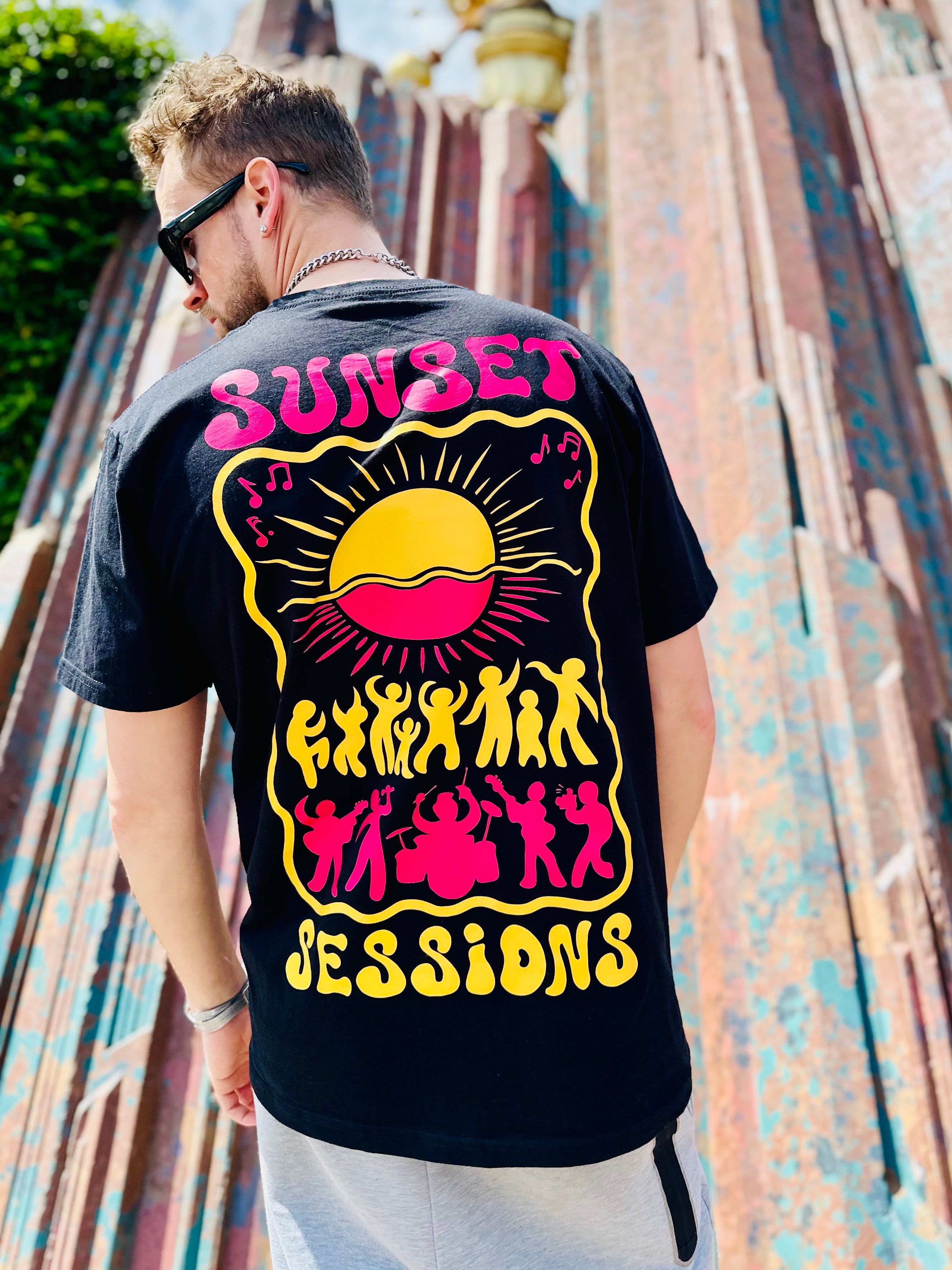 Person wearing a black t-shirt with 'Sunset Sessions' design on the back, standing against a colorful wall.