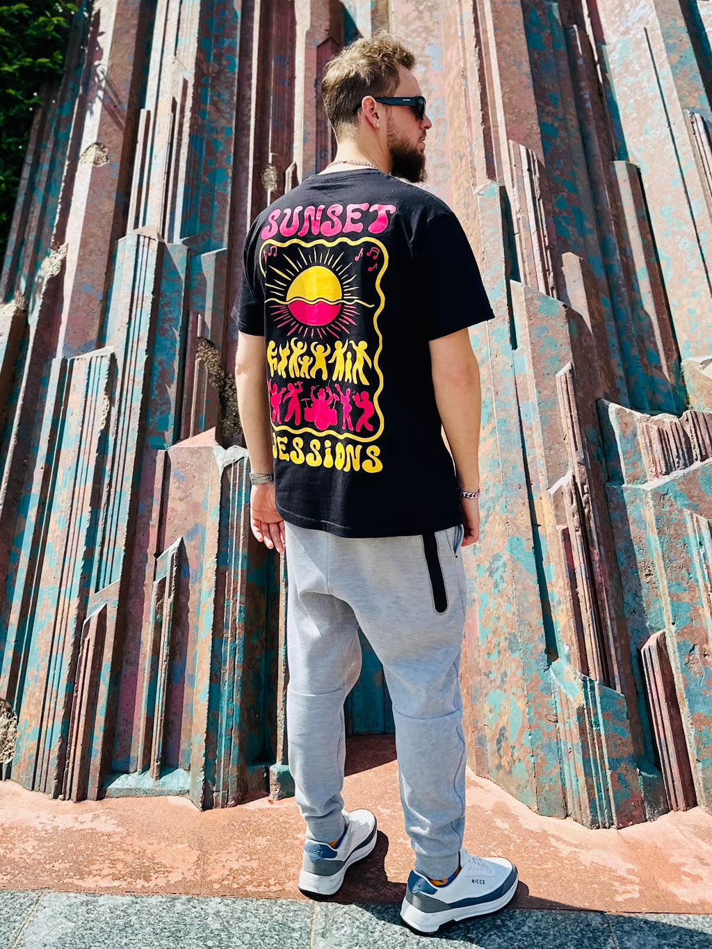 Person wearing a black t-shirt with colorful graphics and text, standing in front of a textured wall.