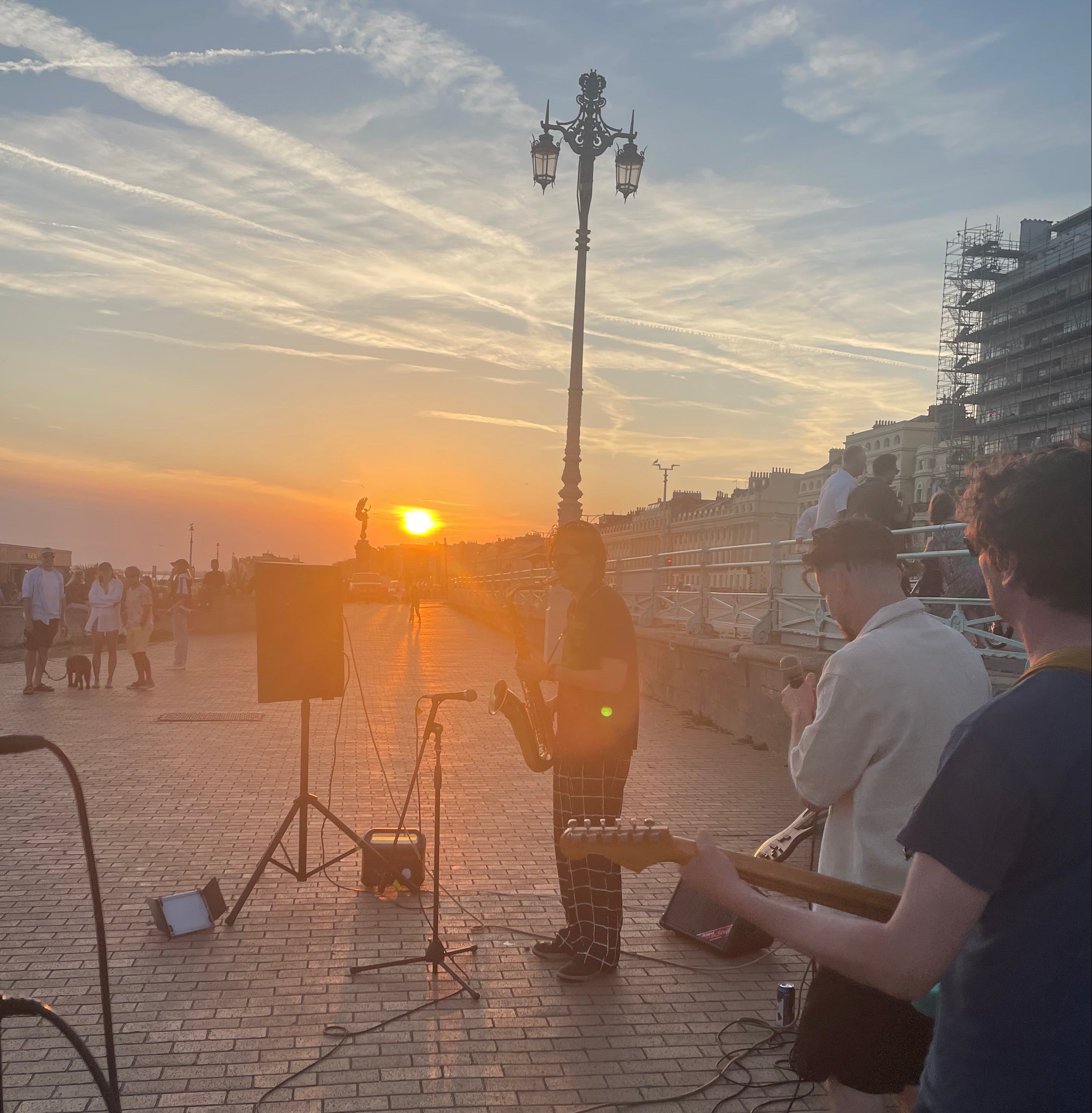Two people playing guitar on a beach at sunset with a city skyline in the background.